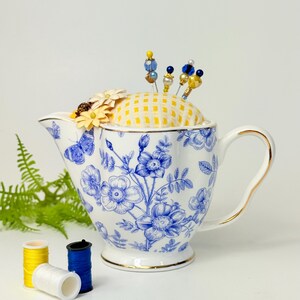 May include: A white and blue floral teapot-shaped pin cushion with a yellow gingham top, holding decorative pins. The teapot has a gold trim and handle. Three spools of thread in yellow, white, and blue are in the foreground.