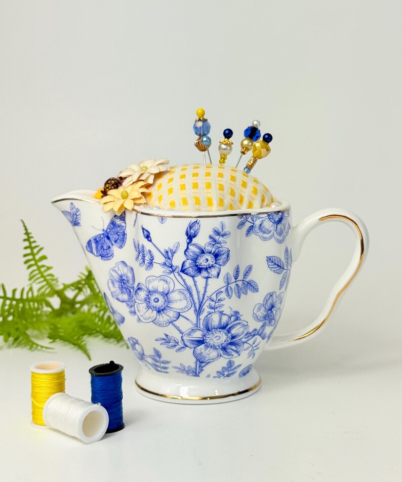 May include: A white and blue floral teapot-shaped pin cushion with a yellow gingham top, holding decorative pins. The teapot has a gold trim and handle. Three spools of thread in yellow, white, and blue are in the foreground.