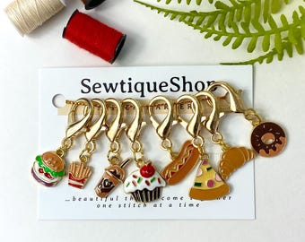 May include: Set of seven gold-tone stitch markers with different food charms. The charms include a burger, fries, a milkshake, a cupcake, a hot dog, a slice of pizza, and a donut. The stitch markers are on a white card with the text "SewtiqueShop... beautiful things come one stitch at a time".