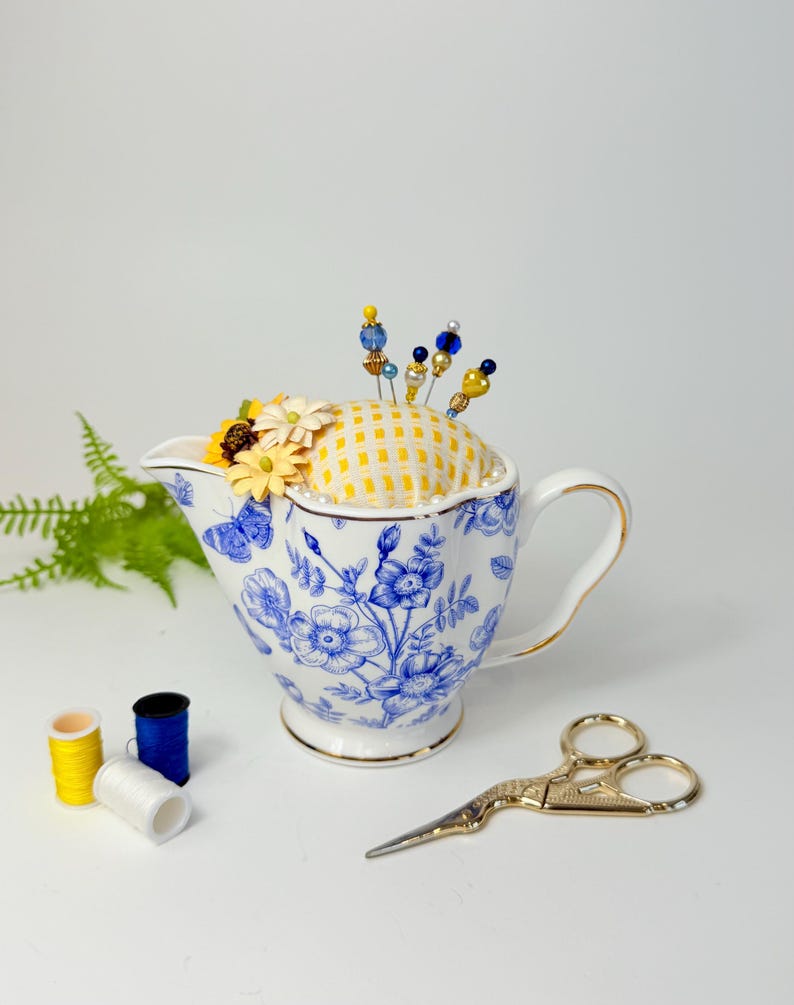 Handmade repurposed pincushion made from a cobalt blue and white creamer pitcher. The cushion is crafted from floral fabric and is tucked neatly into the pitcher’s opening. Embellished with subtle trim and includes decorative pins. Ideal as a sewing supply, quilting gift, or cottagecore room accent.