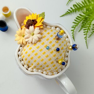 May include: A white ceramic pin cushion shaped like a teacup, filled with a yellow patterned fabric. The cushion is adorned with pearl beads, decorative pins, and fabric flowers in yellow and cream colors. A charming sewing accessory.