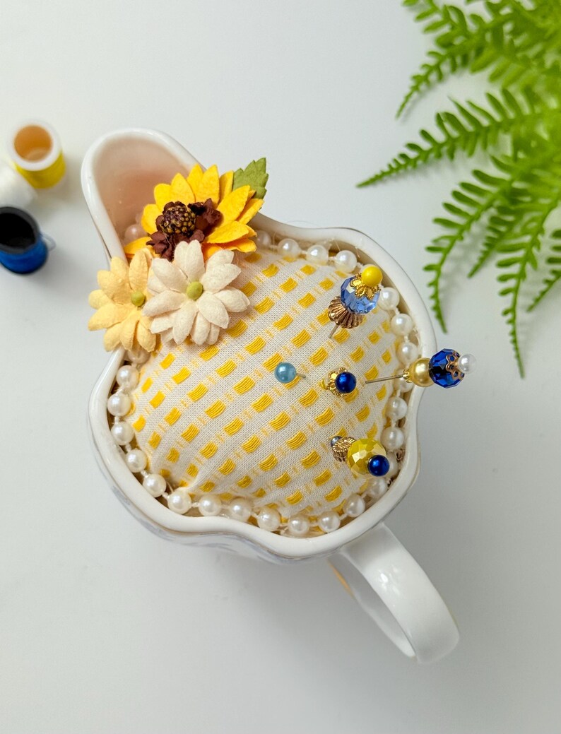 May include: A white ceramic pin cushion shaped like a teacup, filled with a yellow patterned fabric. The cushion is adorned with pearl beads, decorative pins, and fabric flowers in yellow and cream colors. A charming sewing accessory.