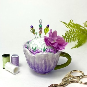 May include: A purple ceramic teacup pincushion with a white floral design and a pink flower on top. The pincushion has green and purple beads on the pins. There are two spools of thread and a pair of gold scissors on a white surface.