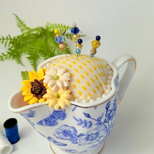 May include: A decorative pin cushion in a white and blue floral gravy boat. The cushion is yellow with a grid pattern and adorned with decorative pins with beaded tops. Artificial flowers and faux pearls add to the charm.
