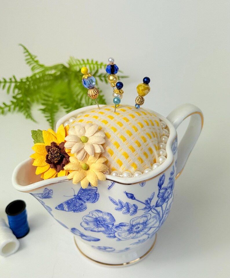 May include: A decorative pin cushion in a white and blue floral gravy boat. The cushion is yellow with a grid pattern and adorned with decorative pins with beaded tops. Artificial flowers and faux pearls add to the charm.