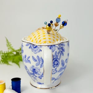 May include: A decorative pin cushion in a teacup. The teacup is white with blue floral patterns and a gold rim. The cushion is yellow with a grid pattern and adorned with beaded pins in various colors. Sewing notions are in the background.