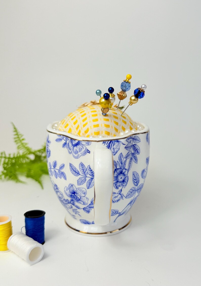 May include: A decorative pin cushion in a teacup. The teacup is white with blue floral patterns and a gold rim. The cushion is yellow with a grid pattern and adorned with beaded pins in various colors. Sewing notions are in the background.