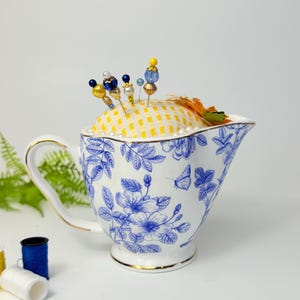 May include: A white and blue floral patterned teacup with a yellow checkered pincushion. Decorative pins with colorful beads are inserted into the pincushion. The teacup has a gold rim and handle. Sewing notions are in the background.