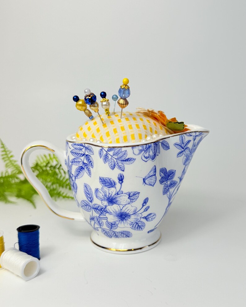 May include: A white and blue floral patterned teacup with a yellow checkered pincushion. Decorative pins with colorful beads are inserted into the pincushion. The teacup has a gold rim and handle. Sewing notions are in the background.