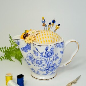 May include: A white and blue floral patterned teacup with a yellow and white checkered pincushion. Decorative pins with beaded tops are inserted into the pincushion. Yellow and white flowers are placed on the pincushion. Sewing supplies are in the foreground.