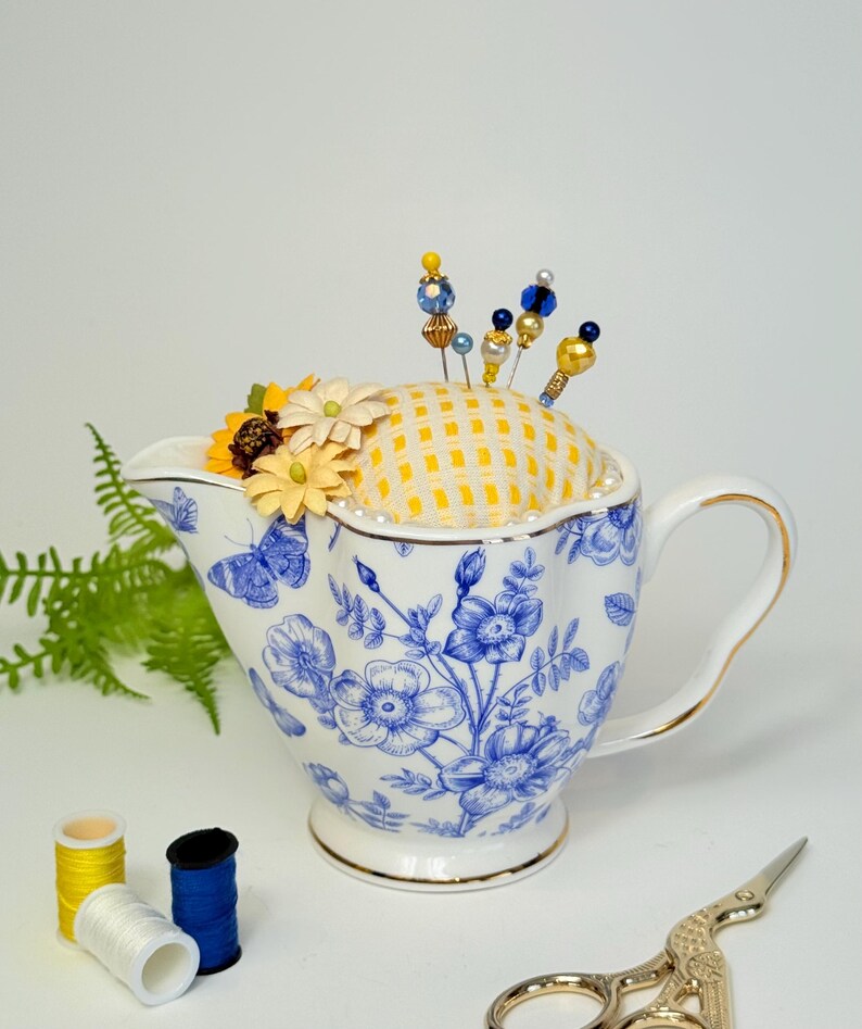 May include: A white and blue floral patterned teacup with a yellow and white checkered pincushion. Decorative pins with beaded tops are inserted into the pincushion. Yellow and white flowers are placed on the pincushion. Sewing supplies are in the foreground.