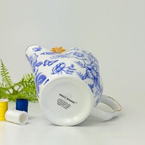 May include: White porcelain creamer with blue floral patterns and a small orange flower accent. The creamer has a handle with gold trim and the text "GRACE TEAWARE" on the bottom. The creamer is made in China.