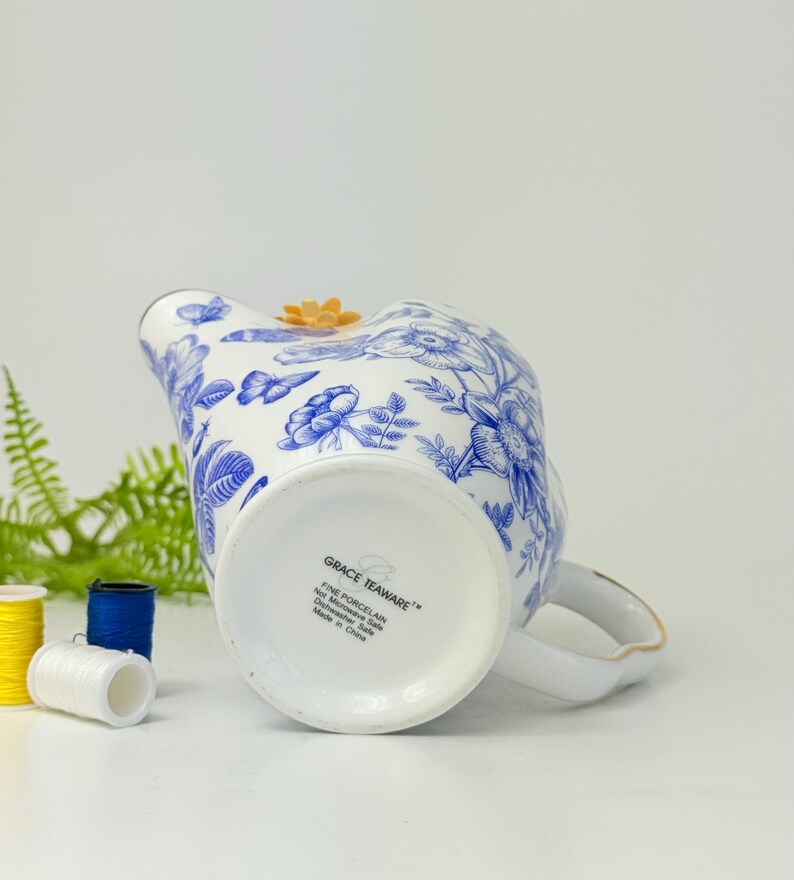 May include: White porcelain creamer with blue floral patterns and a small orange flower accent. The creamer has a handle with gold trim and the text "GRACE TEAWARE" on the bottom. The creamer is made in China.