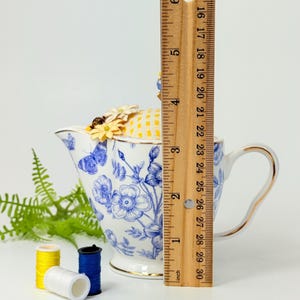 May include: A white and blue floral teapot-shaped pin cushion with a yellow and white checkered top, adorned with small yellow flowers. The teapot measures approximately 6 inches in height. Three spools of thread in yellow, white, and blue are in the foreground.