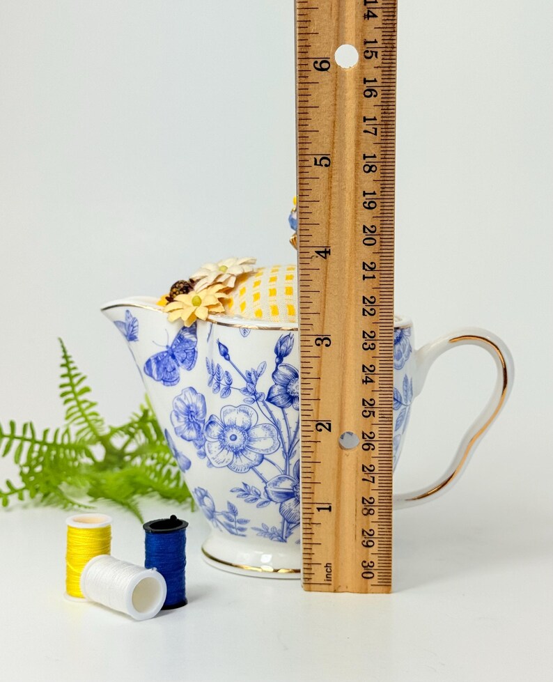 May include: A white and blue floral teapot-shaped pin cushion with a yellow and white checkered top, adorned with small yellow flowers. The teapot measures approximately 6 inches in height. Three spools of thread in yellow, white, and blue are in the foreground.