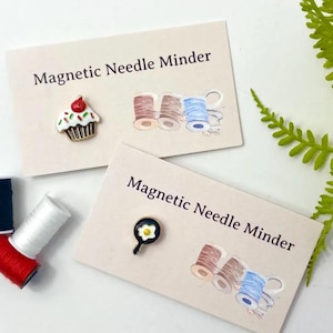 Könnte beinhalten: Zwei Karten mit dem Text "Magnetic Needle Minder" und Bildern eines Cupcakes und einer Bratpfanne mit einem Spiegelei. Die Karten liegen auf einer weißen Oberfläche mit Garnrollen.