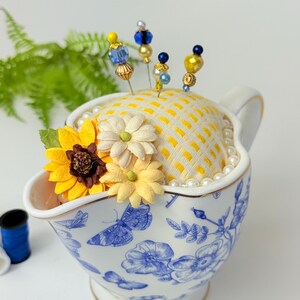 May include: A decorative pin cushion in a white and blue floral patterned gravy boat. The cushion is yellow with a grid pattern and adorned with decorative pins with beaded tops. Artificial sunflowers and pearls add to the design.