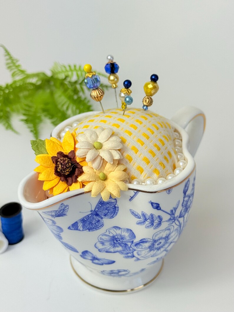 May include: A decorative pin cushion in a white and blue floral patterned gravy boat. The cushion is yellow with a grid pattern and adorned with decorative pins with beaded tops. Artificial sunflowers and pearls add to the design.