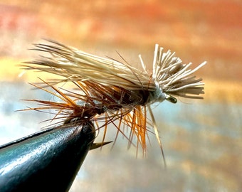 May include: A close-up of a brown and tan fly fishing lure with a gold hook. The lure is tied with feathers and fur.