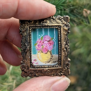 May include: A small painting of pink roses in a yellow vase, set against a teal background with white vertical stripes. The artwork is framed in an ornate, antique gold-coloured frame. The painting is held between two fingers.