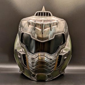 Puede incluir: Un casco metálico verde y plateado con una visera oscura. El casco tiene una superficie texturizada con diseños angulares y una parte superior puntiaguda. El diseño sugiere un tema futurista o de ciencia ficción.