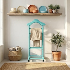 Peut inclure: Un portant bleu turquoise avec un pantalon beige suspendu. Un panier en osier avec des serviettes roulées est posé au sol. Un petit olivier en pot et une étagère en bois avec des plats et des plantes sont également visibles.