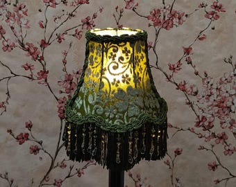 May include: A decorative lampshade with a green floral pattern and beaded fringe. The lampshade has a gold interior and is trimmed with green trim. The background features a floral wallpaper.