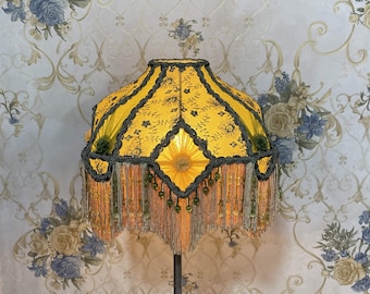 May include: A yellow lampshade with black and green trim and fringe. The lampshade has a floral pattern and is decorated with beads.