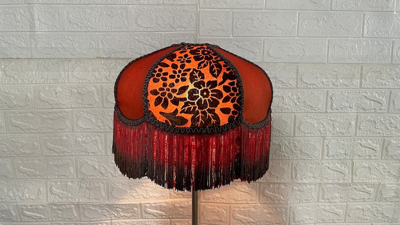 May include: A red and orange lampshade with a floral pattern and a black fringe trim. The lampshade is made of fabric and has a beaded fringe.