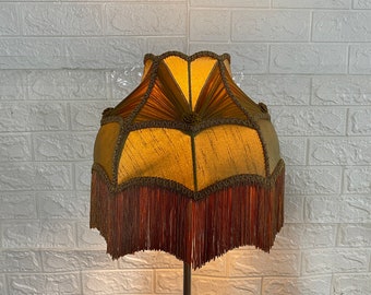 Op de afbeelding: Een lampenkap in vintage stijl met een gouden en bruine stoffen buitenkant. De kap heeft een gegolfde rand en een franje rand.