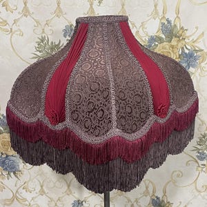 Puede incluir: Una pantalla de l&aacute;mpara de color burdeos y marr&oacute;n con un borde festoneado y ribete de flecos. La pantalla tiene un patr&oacute;n floral y est&aacute; hecha de tela.