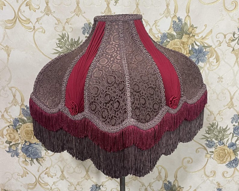 Puede incluir: Una pantalla de l&aacute;mpara de color burdeos y marr&oacute;n con un borde festoneado y ribete de flecos. La pantalla tiene un patr&oacute;n floral y est&aacute; hecha de tela.