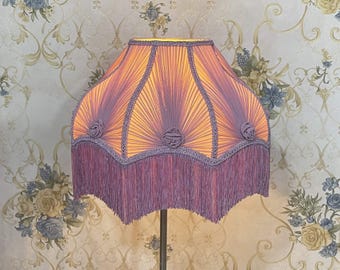 Puede incluir: Una pantalla de lámpara vintage con un diseño plisado en tonos morados y naranjas. La pantalla de la lámpara presenta rosetas decorativas y un ribete de flecos. La lámpara está sobre un fondo de papel pintado floral.