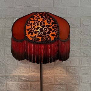 May include: A brown and orange lampshade with a floral pattern and a red fringe. The lampshade is on a black metal stand.