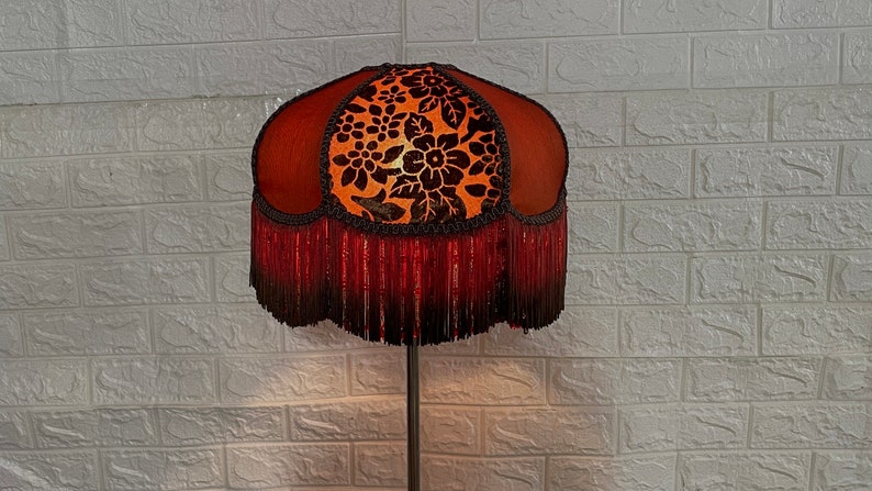 May include: A brown and orange lampshade with a floral pattern and a red fringe. The lampshade is on a black metal stand.
