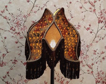 Op de afbeelding: Een decoratieve lampenkap met een uniek ontwerp. De kap heeft een patroonstof in bruin-, oranje- en geeltinten, afgewerkt met donkerbruine franjes en gouden accenten. De lampenkap is ongeveer 61 cm hoog.