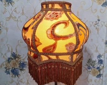 Op de afbeelding: Een gele lampenkap met een bruin en gouden paisley-patroon en franjes. De lampenkap heeft een gegolfde rand en is gemaakt van stof.