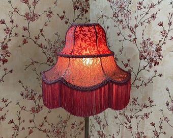 May include: A red lampshade with a floral pattern and fringe trim. The lampshade has a scalloped edge and is lit from within, casting a warm glow. The background features a floral wallpaper.