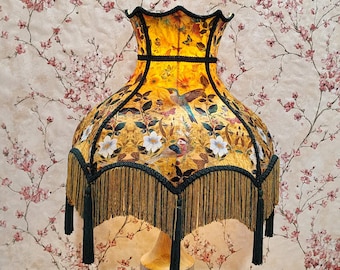 Può includere: Una lampada da tavolo decorativa con paralume giallo a fantasia floreale. Il paralume presenta un disegno di uccelli e farfalle con fiori bianchi e foglie verdi. Il paralume è rifinito con finiture e frange verde scuro.
