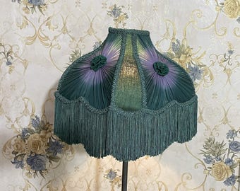 Puede incluir: Una pantalla de lámpara verde con ribete de flecos y detalles florales en morado y verde.