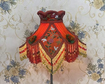 May include: An ornate, orange lampshade with intricate embroidery and beaded fringe. The shade features floral patterns and a decorative trim, creating a vintage aesthetic. The lamp is a statement piece for home decor.