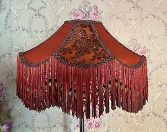 Op de afbeelding: Decoratieve lampenkap met een roestkleurige stof en een bloemmotief. De lampenkap heeft een franjebies met kralenaccenten. Een uniek woondecoratie-item.