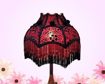 May include: A lampshade featuring a vibrant red fabric base overlaid with a black floral lace pattern. The shade is accented with black braided trim, small black fabric rosettes, and a long black fringe border with dangling black beads.