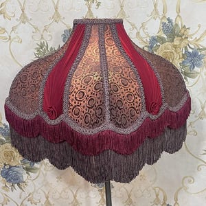 Puede incluir: Una pantalla de l&aacute;mpara de color burdeos y dorado con un borde festoneado y flecos. La pantalla tiene un patr&oacute;n floral y est&aacute; hecha de tela.