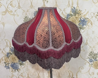 Puede incluir: Una pantalla de lámpara de color burdeos y dorado con un borde festoneado y flecos. La pantalla tiene un patrón floral y está hecha de tela.