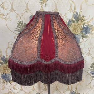 Puede incluir: Una pantalla de l&aacute;mpara roja y dorada con ribete de flecos. La pantalla tiene un dise&ntilde;o plisado y un patr&oacute;n floral.