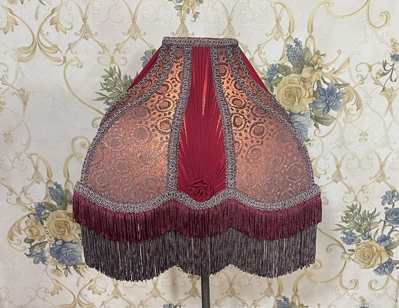 Puede incluir: Una pantalla de l&aacute;mpara roja y dorada con ribete de flecos. La pantalla tiene un dise&ntilde;o plisado y un patr&oacute;n floral.