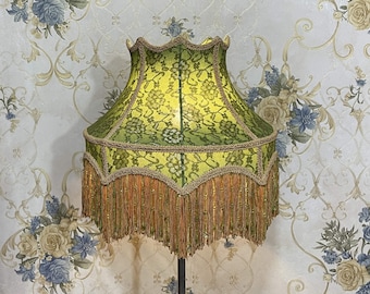 May include: A green and gold lampshade with a lace pattern and fringe trim. The lampshade is made of fabric and has a scalloped edge.