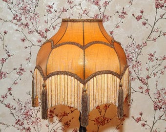 May include: A vintage-style floor lamp with a large, amber-colored lampshade. The shade has a scalloped edge, decorative trim, and a fringe of cream-colored tassels. The lamp is set against a floral wallpaper background.