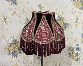 May include: A burgundy lampshade with gold trim and beaded fringe. The shade has a scalloped edge and is decorated with a floral pattern.